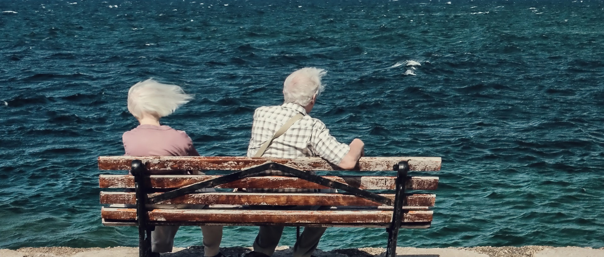 Zu sehen ist ein altes Paar, das auf einer Bank am Meer sitzt und vom Wind durchgepustet wird., © joexx / photocase.deFoto: joexx