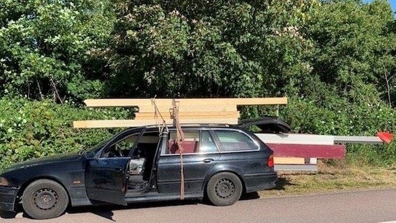 Holzplanken sind auf dem Dach eines PKW mit Schnüren befestigt, weitere Planken reichen aus dem Heck des Autos.  
