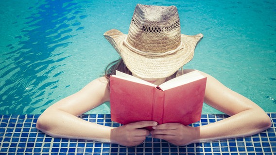 Eine Frau mit einem Sonnenhut steht im Wasser und liest ein Buch. © imago/Westend61 Foto: Westend61