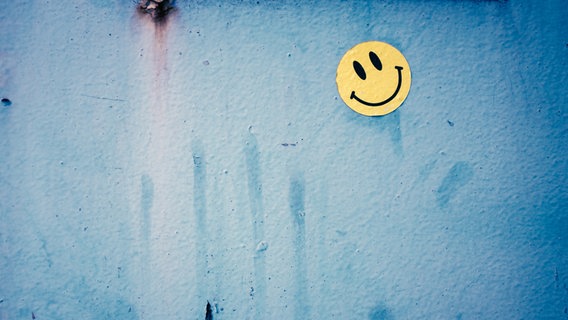 Ein lächelndes gelbes Smiley ist an eine blaue Wand geklebt. © suze / photocase.de Foto: suze / photocase.de