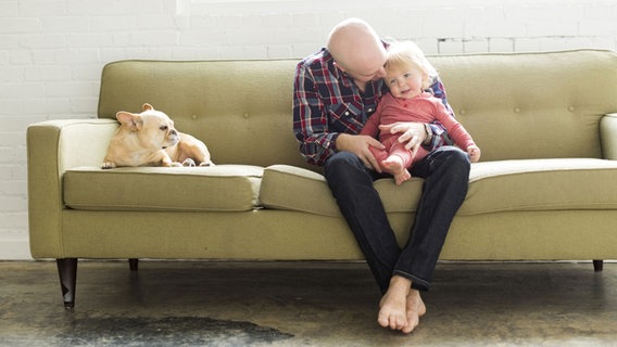 Vater kuschelt mit seinem Baby auf einer grünen Couch. Daneben liegt ein Hund. © picture alliance / Bildagentur-online/Tetra Images Foto: Tetra Images