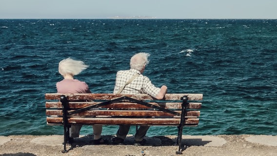 Zu sehen ist ein altes Paar, das auf einer Bank am Meer sitzt und vom Wind durchgepustet wird. © joexx / photocase.de Foto: joexx