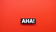 Zu sehen ist der Schriftzug "AHA!" in weißen Buchstaben auf rotem Grund.  