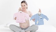 Zu sehen ist eine Frau, die zwei Babyjäckchen hochhält - rosa und blau. © imago / Westend61 