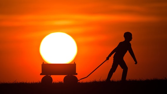 Ein Kind zieht im Sonnenuntergang einen Bollerwagen hinter sich her. Es sieht aus, als würde es die Sonne im Bollerwagen transportieren. © picture-alliance/ dpa - Report Foto: Patrick Pleul