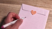 Ein Umschlag mit einem Herz, darunter eine Hand, die einen Stift hält. © pontchen / photocase.de Foto: pontchen / photocase.de