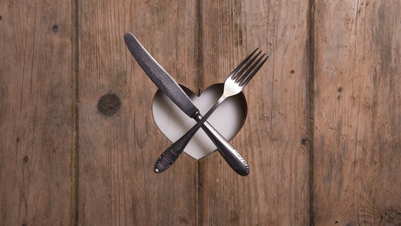 Messer und Gabel liegen überkreuzt auf einem Herz, das in ein Stück Holz gestanzt ist. © arthurbraunstein / photocase.de Foto: arthurbraunstein / photocase.de
