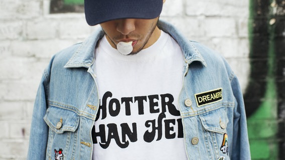 Ein junger Mann mit einer Cappy, einem Kaugummi, einer Lederjacke und einem Shirt mit der Aufschrift "Hotter than hell". © imago/Westend61 Foto: Westend61