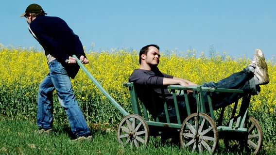 Ein Mann zieht einen anderen Mann in einem altmodischen Karren neben einem Rapsöl-Feld entlang. © photocase.de / dommy Foto: photocase.de / dommy