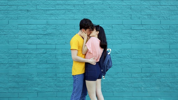 Ein junges Paar küsst sich vor einer blauen Wand. © imago/Westend61 