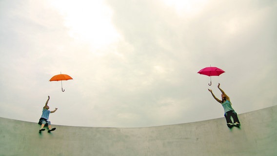 Zwei Personen sitzen auf einer Mauer und greifen nach ihren wegfliegenden Regenschirmen. © Mr. Nico / photocase.de Foto: Mr. Nico