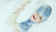 Eine junge Frau mit blauem Haar und rotem Lippenstift schläft in weißer Bettwäsche. © nanihta / photocase.de Foto: nanihta