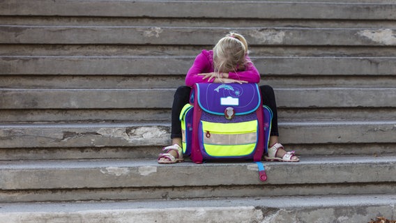 Ein trauriges Kind sitzt, auf seinen Schulranzen gelehnt, auf einer Treppe. © imago/Westend61 
