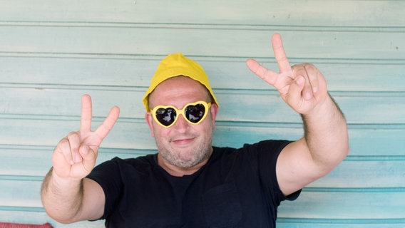 Ein Mann mit einer gelben Mütze und einer Herzchen-Sonnenbrille macht Peace-Zeichen in die Kamera. © suschaa / photocase.de Foto: suschaa / photocase.de