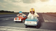 Zwei Jungs mit Helmen fahren in kleinen Autos ein Rennen. © imago/Westend61 Foto: Westend61