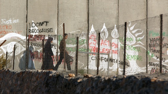 Eine Familie geht zwischen einem Zaun und der Mauer entlang. © NDR Foto: Simon Kremer