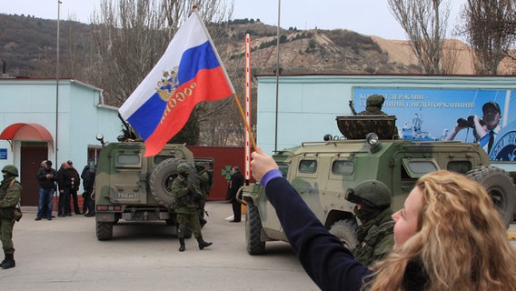 Eine Frau schwenkt in Balaklava eine russische Fahne. Im Hintergrund sind Soldaten zu sehen. © dpa Foto:  ITAR-TASS / Alexei Pavlishak)