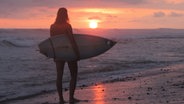 N-JOY Moderatorin Isa Scholz mit einem Surfbrett am Strand bei Sonnenuntergang. © Isa Scholz 