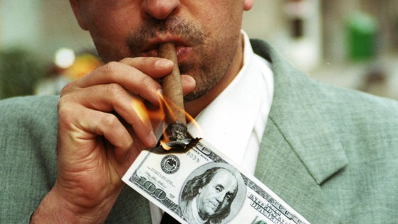 Das Bild zeigt einen Anzugträger, der sich mit einer 100-Dollar-Note eine Zigarre anzündet. © Imago / imagebroker / Rosseforp Foto: Imago / imagebroker / Rosseforp